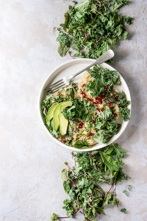 Vegan quinoa salad with kale, young beetroot leaves, garnet seeds, sliced avocado in white plate with ingredients above over grey texture background. Top view, space. Healthy eatingの写真素材