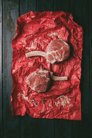 Raw uncooked black angus beef tomahawk steaks on bones served with salt pepper on red crumpled paper over dark wooden plank background. Top view, space.の写真素材