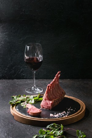 Grilled sliced rack of lamb with yogurt mint sauce served with green salad young beetroot leaves, glass of red wine, pink salt on round wooden slate board over dark black table background. Copy spaceの写真素材