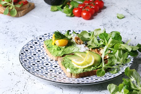 Vegetarian sandwiches with avocado, ricotta, egg yolk, spinach, cherry tomatoes on whole grain toast bread on ceramic plate with ingredients above over white marble kitchen table. Close upの写真素材