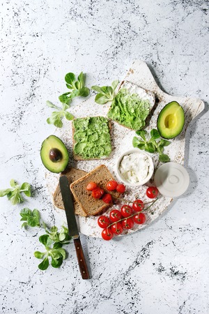 Vegetarian sandwiches with avocado, ricotta, egg yolk, spinach, cherry tomatoes on whole grain toast bread on white serving board with ingredients above over white marble background. Top view, spaceの写真素材