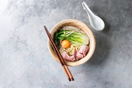 Asian dish udon noodles with egg yolk, sesame, mushrooms, boc choy, sliced sous vide cooked meat served in ceramic bowl with spoon and chopsticks over grey texture background. Top view, space.の写真素材