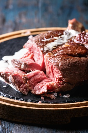 Sliced grilled sous-vide black angus beef tomahawk steak on bone served with salt, pepper, rosemary and white sauce on round wooden slate cutting board over dark wooden plank background. Close upの写真素材
