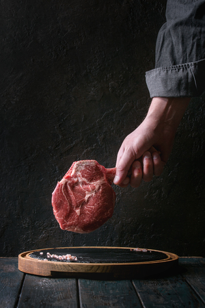 Man's hands holding raw uncooked black angus beef tomahawk steak on bone with salt and pepper on round wooden slate cutting board over dark wooden plank table. Rustic styleの写真素材