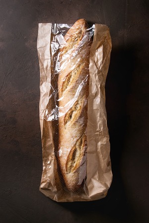 Loaf of fresh baked artisan baguette bread in market paper bag over dark brown texture background. Top view, copy space.の写真素材