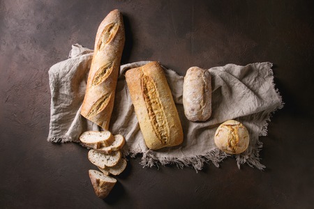 Variety of loafs fresh baked artisan white and whole grain bread on linen cloth over dark brown texture background. Top view, copy space.の写真素材