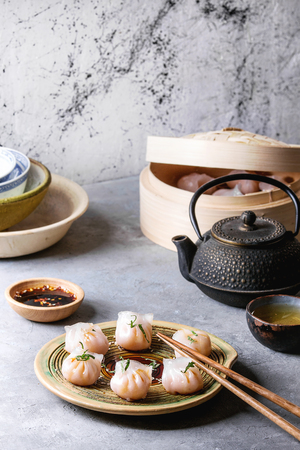 Asian steam potstickers dumplings stuffed by shrimps, served on ceramic plate with soy sesame sauce, chopsticks, teapot, tea, bamboo steamer over grey kitchen table.の写真素材