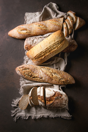 Variety of loafs fresh baked artisan rye, white and whole grain bread on linen cloth over dark brown texture background. Top view, copy space.の写真素材