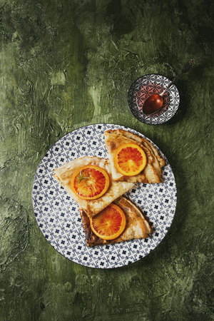 Homemade crepes pancakes served in white decorate ceramic plate with bloody oranges and rosemary syrup with sliced sicilian red oranges over green texture background. Top view, space. Toned imageの写真素材