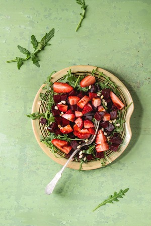 Beetroot and strawberry salad served with arugula and nuts on ceramic plate with fork over green texture background. Top view, space. Healthy eatingの写真素材