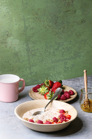 Sweet rice porridge pudding in ceramic plate with berries strawberry and raspberry, walnuts, honey and mug of milk on grey kitchen table with green wall as background.の写真素材