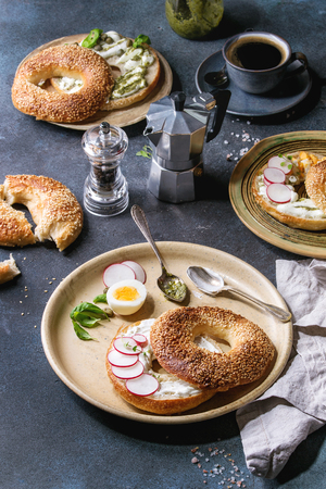 Variety of homemade bagels with sesame seeds, cream cheese, pesto sauce, eggs, radish, herbs served on ceramic plate with ingredients and coffee above over blue texture background. Top view, space.の写真素材