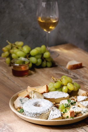 Cheese plate assortment of french cheese served with honey, walnuts, bread and grapes on ceramic plate and glass of white wine over wooden table with grey wall as background.の写真素材