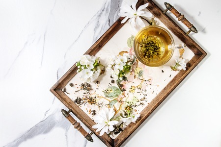 Glass cup of hot green tea on vintage tray with spring flowers white magnolia and cherry blooming branches over white marble texture background. Top view, space.の写真素材