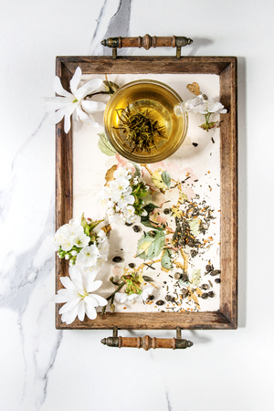 Glass cup of hot green tea on vintage tray with spring flowers white magnolia and cherry blooming branches over white marble texture background. Top view, space.の写真素材