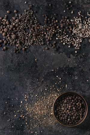 Variety of different black peppers allspice, pimento, monks pepper, peppercorns and ground powder in tin can over old black iron texture surface. Top view, space. Food backgroundの写真素材