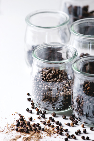 Variety of different black peppers allspice, pimento, long pepper, monks pepper, peppercorns and ground powder in glass jars on white marble kitchen table.の写真素材