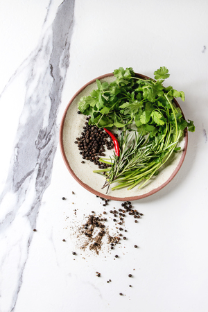 Black pepper peppercorns, ground powder, chili pepper and fresh herbs in ceramic plate over white marble background. Top view, space.の写真素材