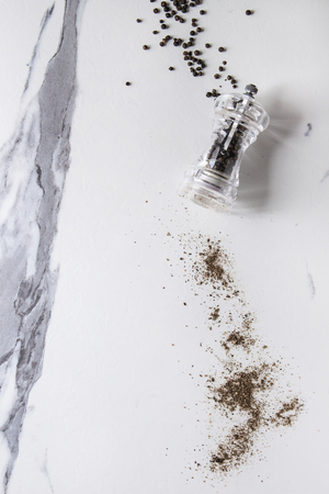 Black peppers peppercorns and ground powder from transparent pepper mill over white marble texture background. Top view, space.の写真素材