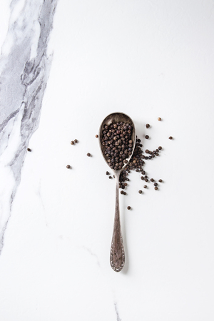 Black pepper peppercorns in vintage spoon over white marble texture background. Top view, space.の写真素材