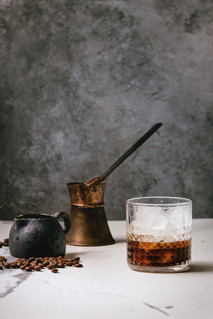 Iced coffee cocktail or frappe with ice cubes in glass with vintage jezva, milk jug and coffee beans around on white marble table with grey concrete wall at background.の写真素材