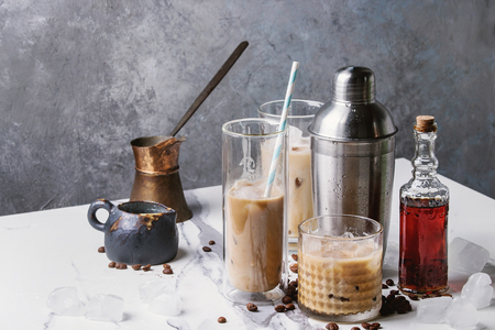 Iced coffee cocktail or frappe with ice cubes and cream in different glasses with jezva, silver shaker, bottle of rum, coffee beans around on white marble table with grey concrete wall at background.の写真素材