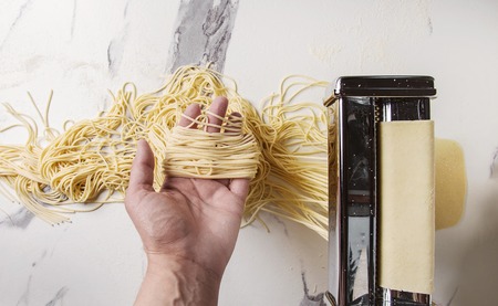 Rolled dough for homemade italian uncooked pasta spaghetti from pasta machine maker in hands with semolina flour over white marble texture background. Flat lay, space.の写真素材
