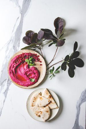 Homemade spread beetroot hummus with pomegranate seeds, olive oil, basil served on ceramic plate with pita bread and green branch over white marble background. Flat lay, space. Mediterranean snack.の写真素材