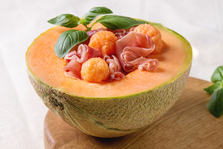 Melon and ham or prosciutto salad served in half of Cantaloupe melon, decorated by fresh basil standing on wooden serving board over white tablecloth. Close upの写真素材