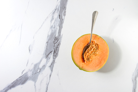Half of ripe organic Cantaloupe melon with seeds and spoon inside over white marble texture background. Flat lay, spaceの写真素材