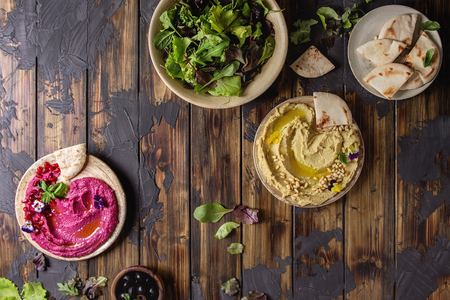Variety of homemade traditional and beetroot spread hummus with pine nuts, olive oil, pomegranate served on ceramic plates with pita bread and green salad on dark wooden background. Flat lay, space.の写真素材