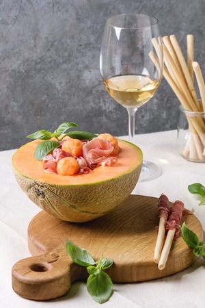 Melon and ham or prosciutto salad served in half of Cantaloupe melon, decorated by fresh basil standing on wooden serving board over white tablecloth with glass of white wine.の写真素材