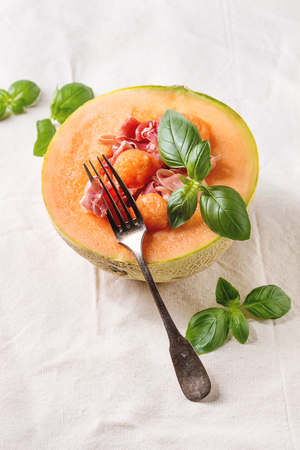 Melon and ham or prosciutto salad served in half of Cantaloupe melon, decorated by fresh basil over white linen cloth.の写真素材