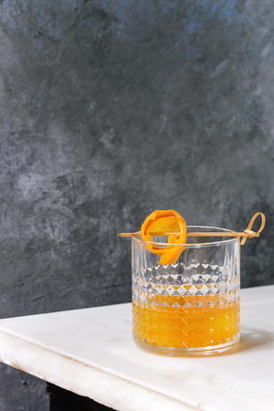 Glass of whiskey orange juice alcohol cocktail with swirled orange peel on skewer standing on white marble table with grey wall at background.の写真素材
