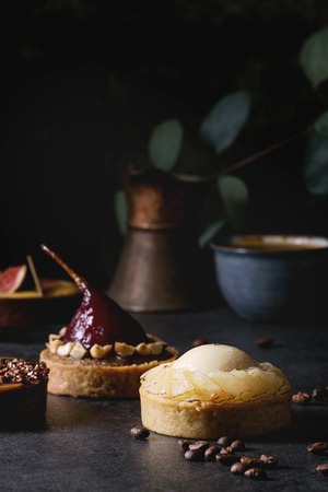 Variety of sweet tartlets with chocolate, caramel, pears, figs with cup of coffee and coffee beans around on black texture table.の写真素材