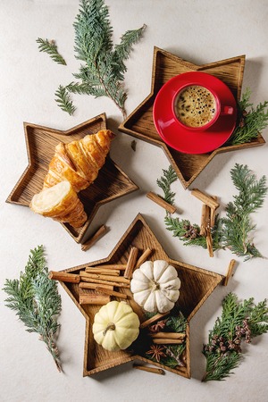 Whole and sliced croissant, red cup of coffee espresso in star shape wooden trays with cinnamon sticks, white decorative pumpkins and thuja branches over white texture background. Flat lay, spaceの写真素材