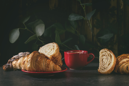 Fresh baked whole and sliced croissant with red cup of coffee espresso on black table with eucalyptus branch. Dark rustic styleの写真素材