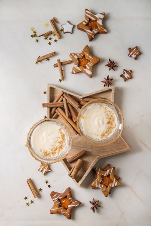 Eggnog Christmas milk cocktail, served in two vintage crystal glasses on wooden tray with shortbread star shape sugar cookies, cinnamon sticks over white marble background. Flat lay, spaceの写真素材