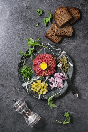 Beef tartare with quail egg in shell, cutting pickled cucumbers, capers, red onion, chives, bread, arugula salad served in marble plates with pepper mill on black texture background. Flat lay, spaceの写真素材