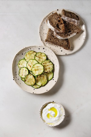 Grilled zucchini salad with yogurt dip and rye sliced bread in spotted ceramic plates over white marble background. Vegetarian food. Flat lay, spaceの写真素材