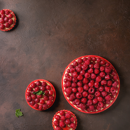 Variety of red raspberry shortbread tarts and tartlets with lemon custard and glazed fresh raspberries over dark brown texture background. Top view, space. Square imageの写真素材