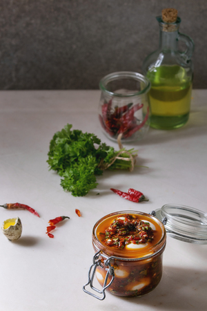 Opened Jar with homemade pickled marinated quail eggs in tomato and olive oil sauce with anchovies and fresh parsley on white marble kitchen table. Ingredients above.の写真素材