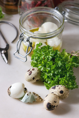 Ingredients for homemade pickled marinated quail eggs. Eggs in plastic box, tomato sauce, chili peppers, anchovies in jars, fresh greens, scissors, olive oil on white marble kitchen table.の写真素材