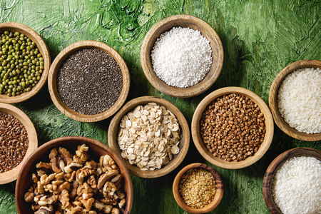 Variety of raw uncooked grains superfood cereal chia linen, sesame, mung bean, walnuts, tapioca, wheat, buckwheat, oatmeal, coconut, rice in wooden bowls over green texture background. Flat lay, spaceの写真素材