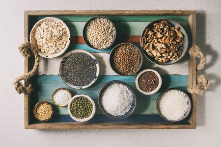 Variety of raw uncooked grains superfood cereal chia seeds, sesame, mung bean, walnuts, tapioca, wheat, buckwheat, oatmeal, coconut, rice in ceramic bowls on wooden plank tray. Flat layの写真素材