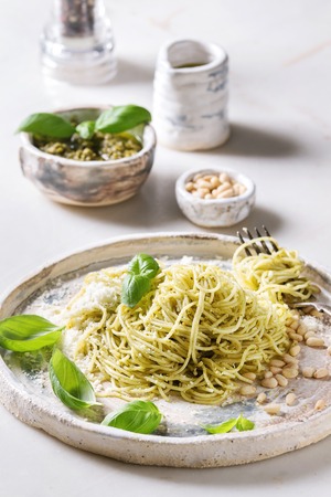 Classic italian spaghetti pasta with pesto sauce, pine nuts, olive oil and fresh basil. Served in ceramic plate with fork and ingredients above over white marble tableの写真素材