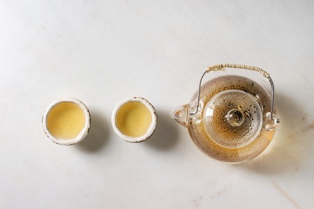 Hot green tea in two traditional chinese clay ceramic cup and glass teapot in row over white marble...