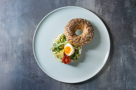 Vegetarian whole grain bagel sandwich with chopped avocado, cream cheese, sun dried tomatoes, egg, served on ceramic plate over blue texture background. Flat lay, spaceの写真素材