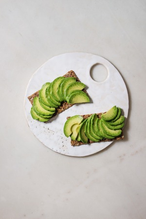 Vegan sandwiches with sliced avocado on rye bread served on ceramic board over white marble...
