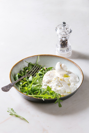 Sliced Italian burrata cheese, fresh arugula salad, pine nuts and olive oil in white ceramic plate on cloth over white marble table.の写真素材